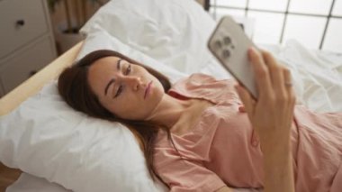 Yatak odasında uzanmış düşünceli bir ifadeyle akıllı telefon ekranına bakan bir kadın.