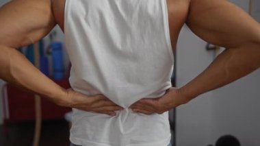 Spor salonundaki genç adam iki eliyle belini tutuyor. Gergin ya da yaralı olduğunu gösteriyor. Beyaz bir atlet giyiyor ve arka planda spor aletleri var.