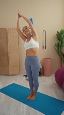Spor salonunda egzersiz yapan bir kadın. Spor malzemeleri ve bitkilerle çevrili yoga minderinin üzerinde geriniyor.