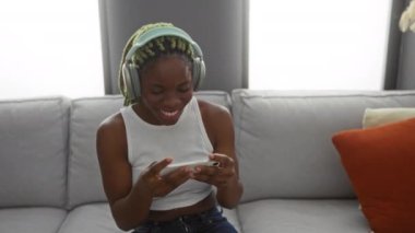 Woman smiling on couch listening to music with headphones and smartphone in cozy living room setting with bright decor.