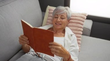 Rahat bir oturma odasında gri bir kanepede dinlenen, gülümseyen bir kitap okuyan, gri saçlarını ve olgun görünümünü sergileyen bir kadın.