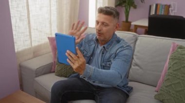 Evde tablet kullanan orta yaşlı bir adam oturma odasındaki kanepede otururken gülümsüyor, video çağrısı yapıyor, günlük kıyafetler, rahat bir atmosfer.