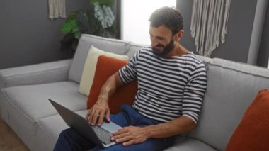 Sakallı genç adam dizüstü bilgisayarından arıyor, gülümsüyor ve modern oturma odasında bitkilerle el sallıyor..