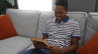 Bir resim çerçevesi tutarken gülümseyen genç adam, modern bir oturma odasında rahat bir kanepeye oturmuş, rahat ve neşeli bir an sergiliyor..