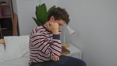 Çizgili tişörtlü genç İspanyol bir adam modern yatak odasında dirseğini tutarak yatağında oturuyor. Arka planda lamba ve bitki var, acı ve rahatsızlık ifade ediyor..
