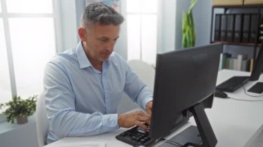 Ofiste çalışan orta yaşlı bir adam bilgisayarlı belgelere odaklanıyor. Modern bir şirket ortamında profesyonel bir çalışma ortamı yaratıyor.