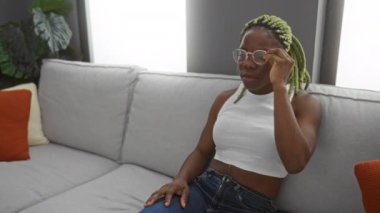 Oturma odasında örgü ören kadın gözlüğünü çıkardıktan sonra yorgun gözlerini ovalıyor, minderli, kapalı bir koltukta yorgunluğunu dile getiriyor, günlük kıyafetler giyiyor ve sakince oturuyor..