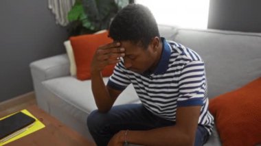 Genç adam modern oturma odasındaki kanepede oturuyor, çizgili gömlek giyiyor, düşünceli ve içe dönük görünüyor, içi rahat, gri kanepe ve turuncu yastıklar var..