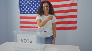 Amerikan bayrağının önünde kapalı bir üniversite odasında oy veren bir kadın, yakınlardaki bir oy sandığıyla işaret ediyor ve gülümsüyor, Amerika Birleşik Devletleri 'ne demokratik katılımı gösteriyor.