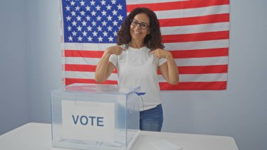 Seçmen odasında kendini gösteren ve arka planda Amerikan bayrağı olan, saydam bir oy sandığının önünde duran, gülümseyen olgun İspanyol bir kadın..