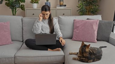 Genç bir İspanyol kadın, bir oturma odasında kanepede oturup dizüstü bilgisayarıyla çalışırken kedisi rahat bir evin içinde yanında uzanıyor..