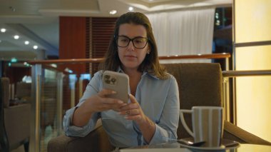 Kadın kahveden zevk alırken, lüks bir bar ortamında telefona bakıyor, genç İspanyol profesyonel yaşam tarzını ve akşam rahatlığının zarafetini vurguluyor..