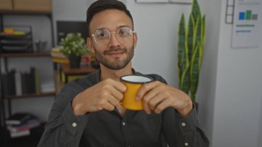 Sakallı, gözlüklü, sarı bardaklı yakışıklı İspanyol bir adam. Arka planda bitki ve raflarla dolu bir ofiste oturuyor..