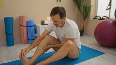 Sağlık merkezinde paspasın üzerinde oturan İspanyol bir adam yoga ekipmanları ve mum ışığıyla çevrili yaralı bacağına masaj yapıyor..