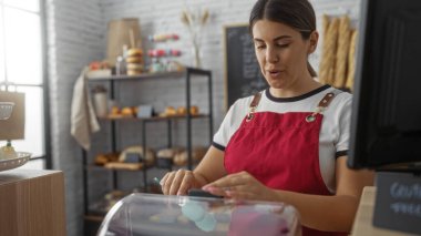 Pastanede çalışan, arka planda çeşitli hamur işleri olan bir kadın. Kapalı dükkan ortamında kırmızı önlük takıyor.