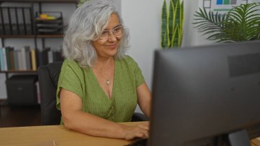 Ofis odasında bilgisayarda çalışan yaşlı bir kadın, iş yerinde kapalı bir ortama dalmış, olgun, gri saçlı, güzel bir kadını resmediyor..