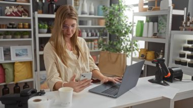 Modern dekorasyon ve ev bitkileriyle çevrili akıllı telefon ve dizüstü bilgisayar kullanarak şık bir ev dekorasyonunda çalışan bir kadın.