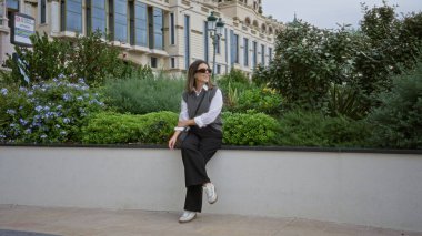 Monaco 'da açık havada gülümseyen bir kadın. Etrafı yemyeşil ve zarif mimari ile çevrili. Huzurlu ve lüks bir atmosferi canlandırıyor..