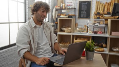 Modern fırında dizüstü bilgisayarda çalışan genç bir adam. Arkaplanda ekmek rafları ve hamur işleri var.