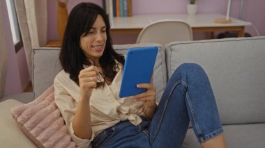 Evindeki oturma odasında dinlenirken tablet okuyan genç bir kadın. Arka planda kitaplar ve pembe bir yastıkla.