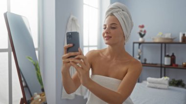 Kaplıcada beyaz bir havlu giyerken telefonuyla selfie çeken genç bir kadın.