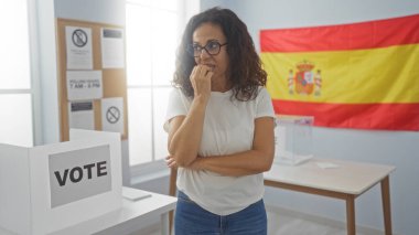 Woman voting indoors at election booth with spanish flag in background, expressing concern during the electoral process