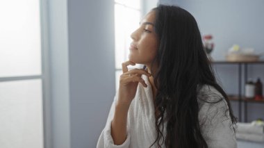 Genç esmer kadın spa merkezinin içinde dinleniyor, beyaz bir bornoz giyiyor ve yumuşak ışıklandırmalı huzurlu bir sağlık odasında kendine hayran.