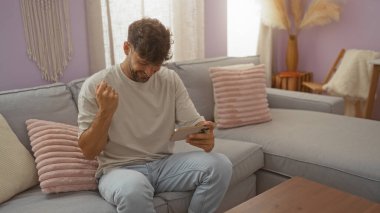 Oturma odasında oturan genç adam elinde bir telefonla heyecanlı görünüyor. Etrafı yastık ve modern dekorlarla çevrili.