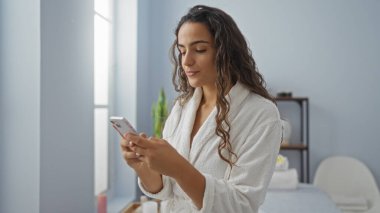 Spa 'daki genç kadın sabahlık giyerken akıllı telefon kullanıyor, rahatlama ve sağlığını iletiyor.