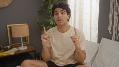 Yakışıklı, genç, İspanyol bir adam sıcak, kapalı bir yatak odasında bir yatakta oturur, sohbet ederken yukarıya doğru işaret eder, rahat ama çekici bir atmosfer yaratır..