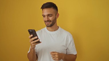 Sakallı yakışıklı, sarı arka planda telefonuna bakarken gülümseyen bir İspanyol..