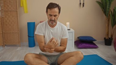 Man holding his wrist in a wellness room with yoga mats and plants, highlighting relaxation and self-care, suggesting a calm spa environment for mature men. clipart