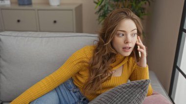 Genç, çekici, sarışın bir kadın rahat bir oturma odasında oturmuş telefonuyla konuşuyor, evde vurulmuş, ev dekorasyonu yapıyor..