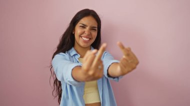 Güzel, genç, İspanyol bir kadın, her iki orta parmağıyla saldırgan bir jest yapıyor. Gülümsüyor, izole pembe bir arka plan duvarına karşı duruyor..