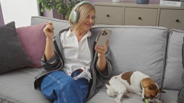 Oturma odasında kulaklıklarla dinlenen olgun sarışın bir kadın telefonuna bakarken ve kanepede küçük köpeğiyle eğlenirken..