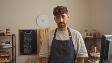 Bir pastanede tezgahın arkasında duran genç adam raflı bir önlük giyiyor ve arka planda fırınlanmış eşyalar ve duvarda bir saat var.