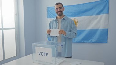 Young hispanic man voting indoors with argentina flag in the background at an electoral college