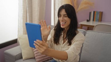 Tablet kullanan ve rahat oturma odasında yastıklar ve kitaplıkla gülümseyen genç bir kadın..