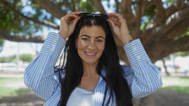 Güzel, genç, İspanyol bir esmer kadın güneşli bir günün tadını çıkarıyor. Parlak bir şekilde gülümsüyor ve arka planda bir bahçe ve yol ile güneş gözlüğünü ayarlıyor..