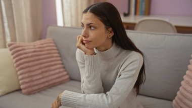 Genç, çekici, esmer, esmer bir kadın bej perdeleri, pembe yastıkları ve arka planda kitapları olan bir oturma odasında düşünceli bir şekilde oturuyor..