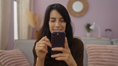 Genç bir kadın pembe aksanlı ve modern dekorlu rahat bir oturma odasında akıllı telefon kullanırken gülümsüyor.