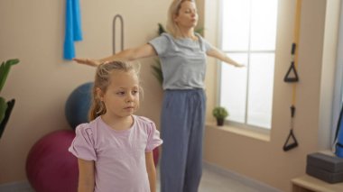 Anne ve kızı spor salonunda birlikte egzersiz yapıyor, sarışın bir kadın ve çocuğu spor salonunda sergiliyor ve spor aletleri etraflarında.