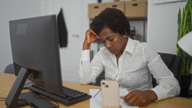 Ofiste çalışan kıvırcık saçlı Afrikalı Amerikalı bir kadın bilgisayar ve akıllı telefonlu bir masada oturmuş modern bir iş ortamını resmediyor..