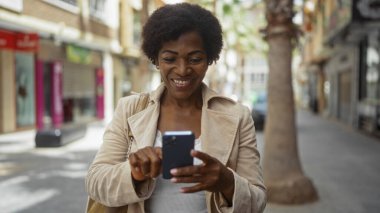 Orta yaşlı kıvırcık saçlı Afrikalı Amerikalı kadın akıllı telefonu şehirde mutlu bir şekilde kullanıyor.