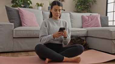 Genç bir İspanyol kadın, oturma odasında yoga minderinde elinde bir kedi, akıllı bir telefon ve odaklanmış bir şekilde oturuyor..