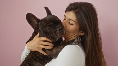 Genç bir İspanyol kadın, pembe arka plan duvarına yaslanmış Fransız bulldog köpeğini sevgiyle kucaklıyor..