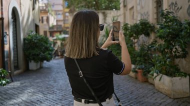 Roma 'nın pitoresk caddesinde canlı yeşilliklerle fotoğraf çeken, İtalyan mimarisini sergileyen ve tarihi eski kasabadaki canlı şehir atmosferini yansıtan bir kadın..