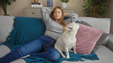 A young, beautiful blonde woman relaxes on a living room sofa with her canine pet indoors, enjoying a peaceful moment together in a cozy apartment. clipart