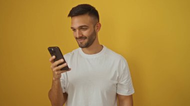 Sakallı yakışıklı genç adam telefonuna bakarken gülümsüyor, beyaz gömlek giyiyor, sarı arka planda soyutlanmış..