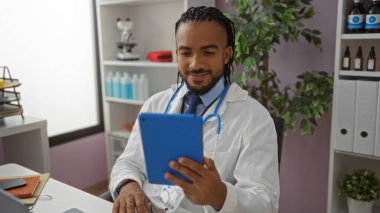 Hastane ofisinde bitki, steteskop ve arka planda rafları olan tablet kullanan beyaz önlüklü bir adam, kapalı alanda odaklanmış ve profesyonel görünüyor..